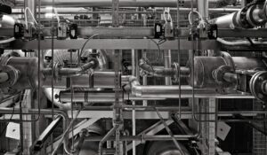 Industrial workers inspect insulated process pipes, valves, gauges, and structural supports inside a facility.