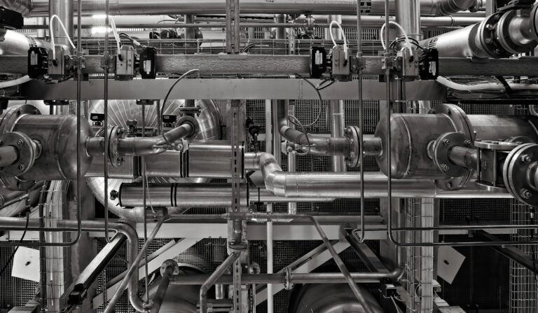 Industrial workers inspect insulated process pipes, valves, gauges, and structural supports inside a facility.